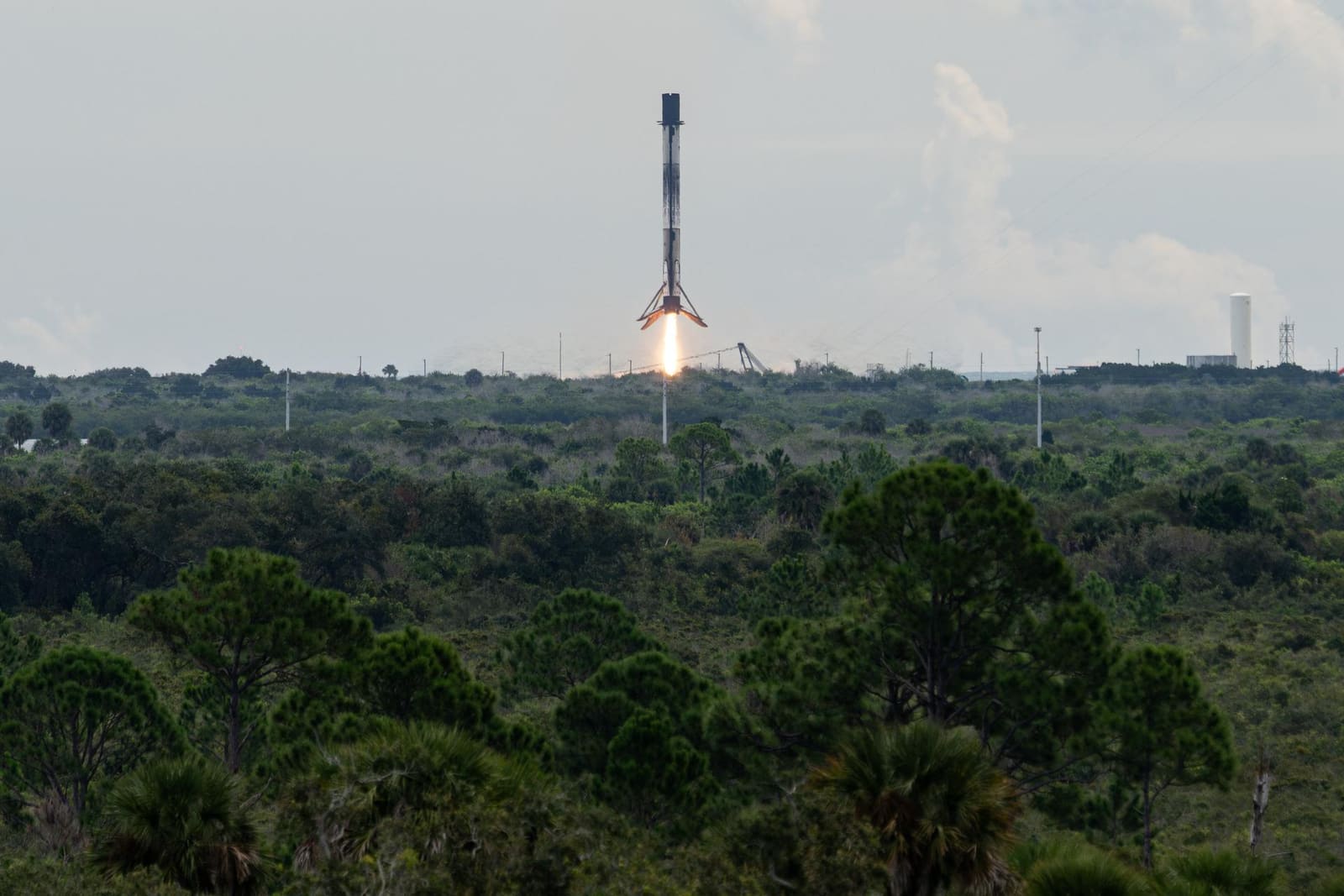 A Falcon 9 first stage descending on a column of flame toward a landing pad, with scrubland in the foreground